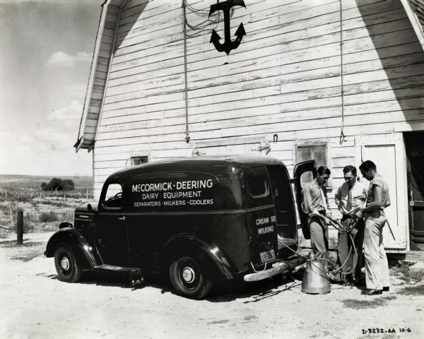 Mr. C.L. Thompson from the Thompson Hardware Company demonstrates a McCormick-Deering milker to an unidentified man and dairyman Wayne Wakefield. The men stand outside of a barn with an anchor symbol on the front near an International vehicle advertising "McCormick-Deering Dairy Equipment, Separators-Milkers-Coolers" on its side.