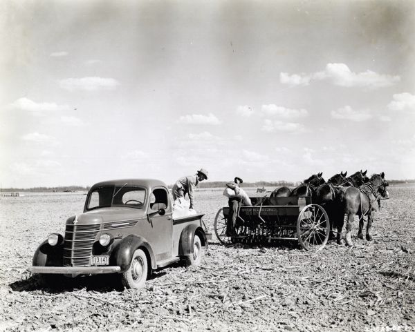 Two men use a McCormick-Deering grain drill with a team of four horses and an International D-2 truck to work in a field on the Purdue University livestock farm. There is a tractor in the background.