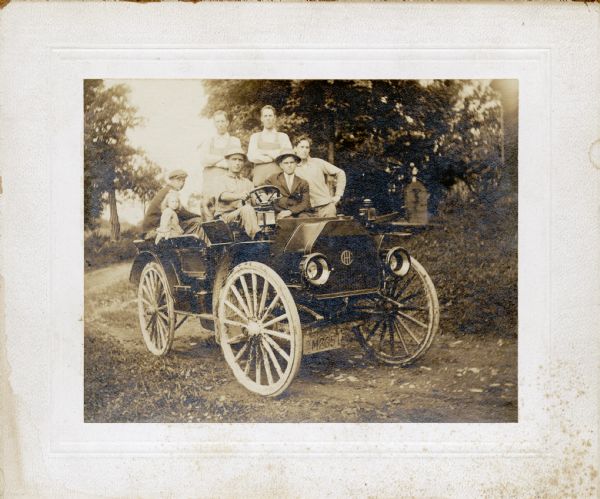Several men and a young girl in an International Auto-Wagon on a dirt road. One or more of the men may have worked for an International dealership. The car has a dealer license plate. An original caption reads: "1915 NY dealer IHC commercial car."
