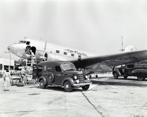 An International U.S. D-15 mail truck used by American Airlines is parked with its rear doors open on a runway in front of an airplane. Two men are working on a metal stairway near the airplane, and a woman is walking towards them. The original caption reads: "Model D-15 panel owned by American Airlines, photograph taken at Chicago Airport by Dell Long, of Kaufmann-Fabry.
