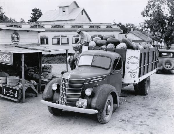 Slightly elevated view of two men loading watermelons into the back of an International D-35 truck owned by the the Redmon Grain Company. Signs advertising Hires R&J Root Beer, Coca-Cola, and Herbola are on the sides of a small wooden stand that holds farm produce. In the background is a railroad car, perhaps parked in the grass, and a tall industrial-style building. The original caption reads: "D-35 owned by the Redmon Grain Co., Hiawatha, Kansas. The load of watermelons shown has been hauled from the Kaw River Valley, a distance of about 120 miles. This truck is used for hauling watermelons about 30 days each summer."