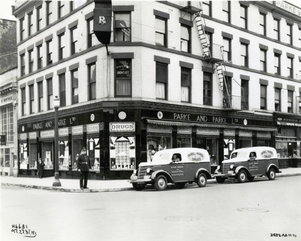 Two men driving International D-2 trucks used by Parke & Parke Drugs park along the road in front of the storefront. A police officer stands on the street corner and a woman exits the store in the background. The original caption reads: "Hamilton, Ontario, Canada. 2 Model D-2, 113" w.b. owned by Parke & Parke druggists."