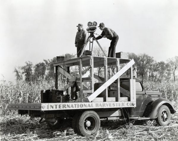 Two men use a platform on the back of an International truck parked in a cornfield to operate a motion film camera. The original caption reads: "Suggested truck platform for motion picture work."