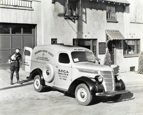 A uniformed man carries a dog to the back of an International D-2 truck used by the Wisconsin Humane Society. The truck is parked in the driveway near the garage door of the Wisconsin Humane Society free animal clinic. There is a sign for the clinic near the doorway of the building. The original caption reads: "D-2 Panel." The text on the truck reads: "Be a humane driver; Don't harm pets or children."
