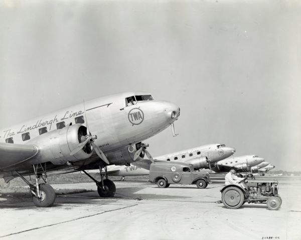 A man uses an International I-12 tractor to move a TWA plane marked: "The Lindbergh Line" along a tarmac. A man is looking out of the window of the plane being moved. An International D-15 truck marked "TWA" and additional planes are in the background. The original caption reads: "Model I-12 tractor and Model D-15 panel owned by TWA, taken at Chicago Airport by Dell Long, Kaufmann-Fabry."