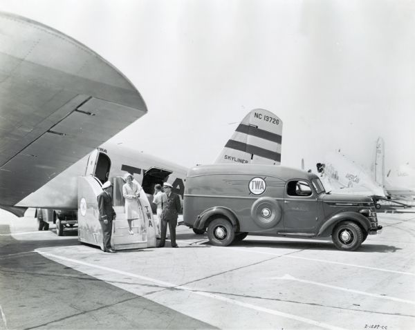 A flight attendant and pilots stand at the portable setps to a TWA plane. An International D-15 truck used by TWA is parked nearby. The original caption reads: "Model D-15 panel owned by TWA, taken by Dell Long, Kaufmann-Fabry."