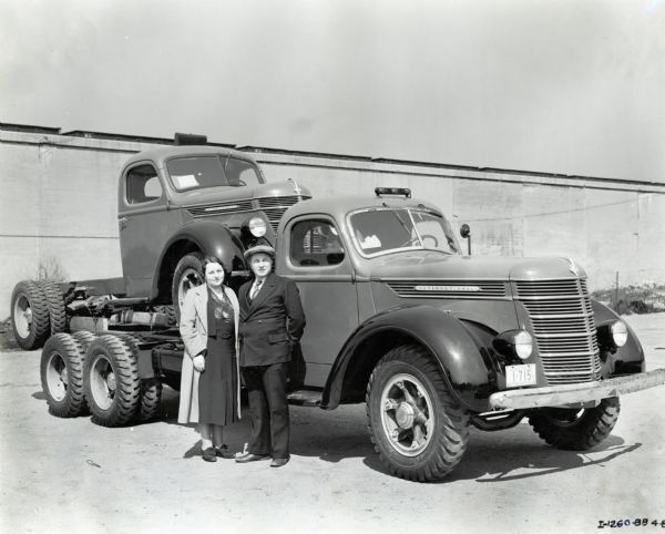 A man and woman are standing in front of two International trucks. One truck is loaded onto the back of the other. The original caption reads: "Bluffton, Alberta; Model D-24 b-F & D-30. Owned by Chas. Montalbetti. Shown at 81st St. Works. Lady is Mrs. Montalbetti."
