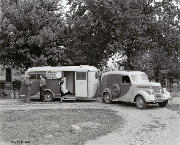 Two men speak to one another as a woman wearing an apron enters a trailer used by the Ohio Edison Company. The trailer is attached to an International D-2 truck and is parked in front of what may be a farmhouse. The original caption reads: "D-2 panel truck and special trailer built by Bender Body Co. This unit is used to sell and demonstrate electrical appliances throughout Ohio."