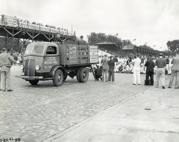 A photographer uses the bed of an International truck parked on the outskirts of a crowd to take photographs at the Indianapolis 500 race. Other photographers are standing near the truck, and a group of people are gathered for the photograph. In the background the grandstand is filled with people. The original caption reads: "D-300 Int. used by the official photographer at the Auto Races." The signs on the side of the truck read: "Easy to Handle, Less Parking Space, Greater Body Length, Ask for Demonstration" and "E.M. Kirkpatrick, Official Photographer. 26th Annual 500 Mile Speedway Race, Indianapolis."