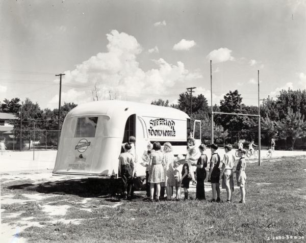 Children line up at the entrance to the Superior Bookmobile trailer.  Other children ride bicycles and play in a playground in the background. The original caption reads: "Superior Bookmobile (traveling library - built by Superior Body Company)." The text on the trailer body reads: "Superior All Steel Bookmobile."