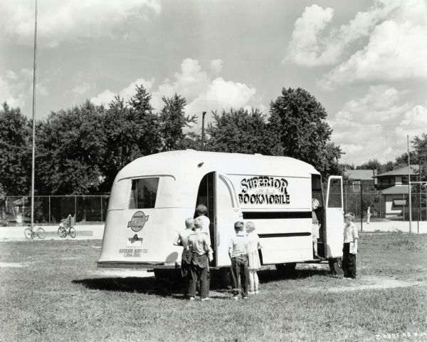 A group of children lines up outside the entrance to the Superior Bookmobile (an International truck), parked in a playground area. Other children holding books exit the trailer. The original caption reads: Superior Bookmobile (traveling library - built by Superior Body Company, Lima, Ohio." The text on the trailer reads: "Superior All Steel Bookmobile."