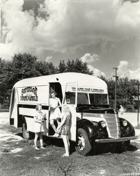 Three girls stand at the entrance of the Superior Bookmobile (an International truck) holding books. A child playing tennis is in the background. The original caption reads: "Superior Bookmobile (Traveling Library - built by Superior Body Company, Lima, Ohio)." The text on the side of the trailer reads: "Superior All Steel Bookmobile."