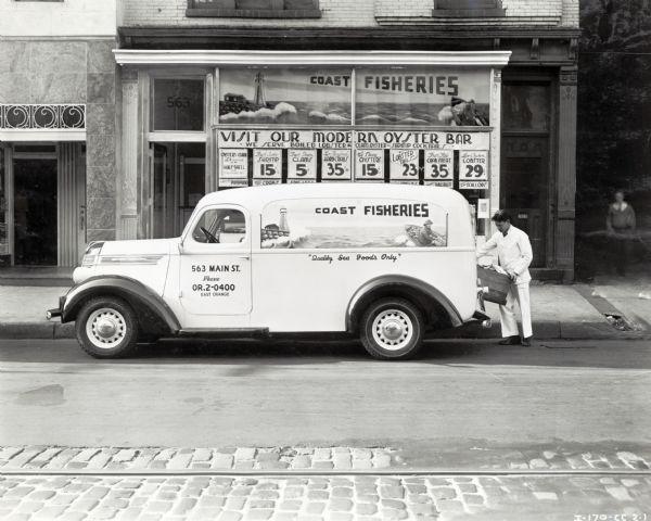 A man loads a wooden crate onto the back of an International D-7  truck used by Coast Fisheries. The truck is parked on the street near the storefront. A man is walking out of the alley on the right. The original caption reads: "D-7 panel. Coast Fisheries." The advertisements in the window read: "Visit Our Modern Oyster Bar; We Serve Boiled Lobster & Clam, Oyster & Shrimp Cocktails." The text on the truck reads: "Quality Sea Foods Only."