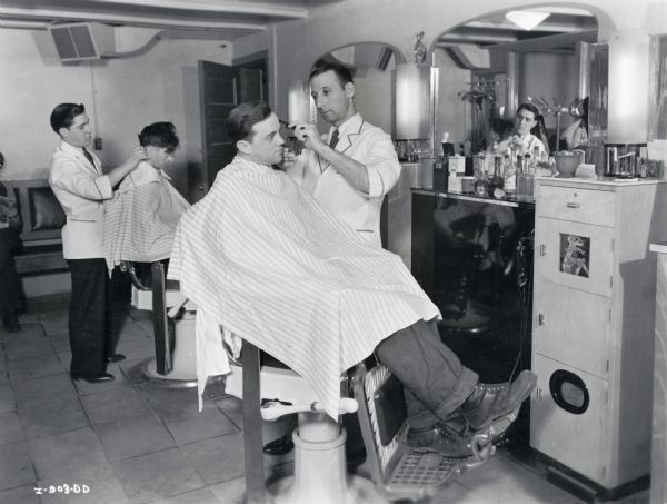 Two men sit in chairs at an American Transporation Company barber shop while barbers clip and comb their hair.