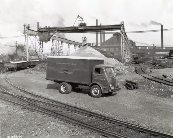 An International D-500 truck owned by the Wickwire Spencer Steel Company is parked in the foreground of an industrial area near railroad tracks. In the background is a crane and industrial buildings with smokestacks. The original caption reads: "D-500 with 361 cu. in. engine, 114" W.B., 14 ft. body, 9:00-10 tires - Wickwire Spencer Steel Co."