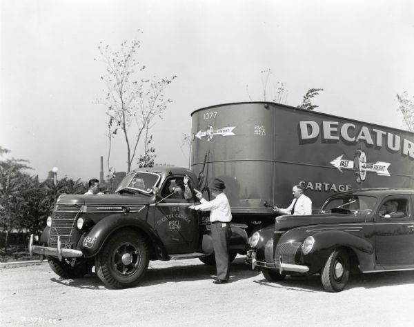 Men hold guns up to the driver of an International D-40 truck marked "Decatur Fast Freight" in a hijacking scene staged by the Chicago Police Department. A man is sitting in the driver's seat of an automobile on the right. The original caption reads: "Highjacking scene staged by members of the Chicago Police Department, Cartage Detail, cooperating with Park District police. The picture was made for possible use in the November, 1939, <i>Trail</i>. The tractor is a Model D-40 owned and operated by the Decatur Cartage Company; Decatur Cartage in addition to supplying the equipment also supplied the driver and an assistant."