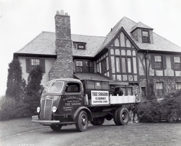 An International D-300 truck owned by P. Hansling & Son Hartford Forestry Company Tree Surgeons is parked in the drive of a home with a large chimney. A man is working near a tree behind the truck. The original caption reads: "Model D-300 owned by P.Hansling & Son, Hartford, Conn. Tree surgeons. This truck is equipped with ventrical-type... immediately behind the cab and hoist which permits its use as a dump body."