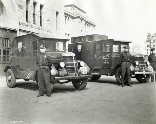 Three uniformed police officers stand beside two parked International trucks marked "Mercer & Dunbar Inc. Armored Car Service." A large building is in the background.