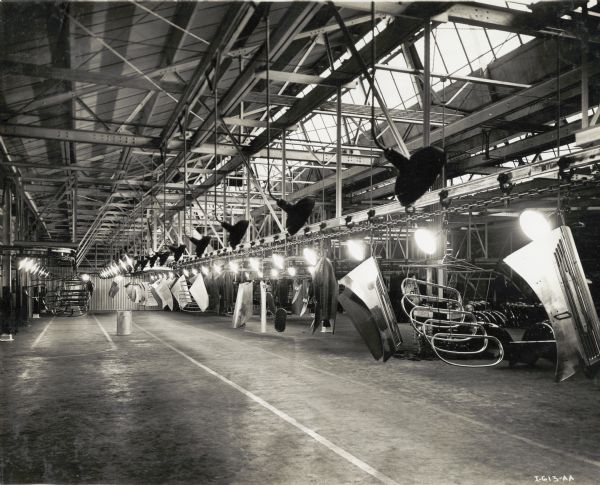 A conveyor system suspended from the ceiling at International Harvester's Springfield Works transports truck parts across the workroom floor. There are skylights in the factory ceiling, and rows of large lamps suspended from the ceiling illuminate the parts.