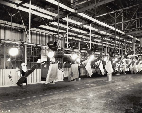 A conveyor system suspended from the ceiling moves truck parts across the floor at International Harvester's Springfield Works factory. Rows of large lamps suspended from the factory ceiling illuminate the parts.