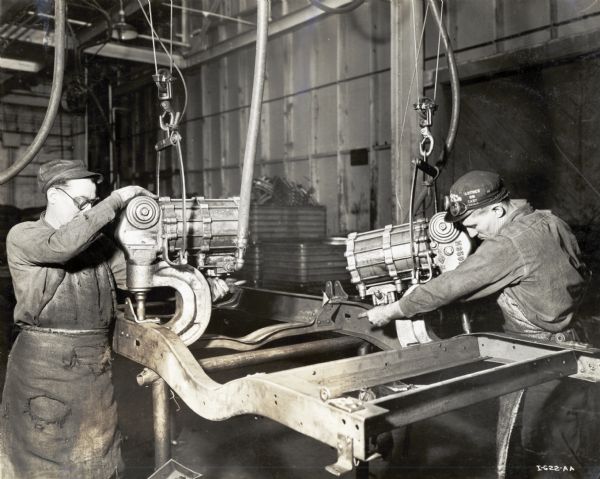 Two men use riveters to construct a truck chassis at International Harvester's Springfield Works factory. The man on the left wears protective eyeglasses and an apron.