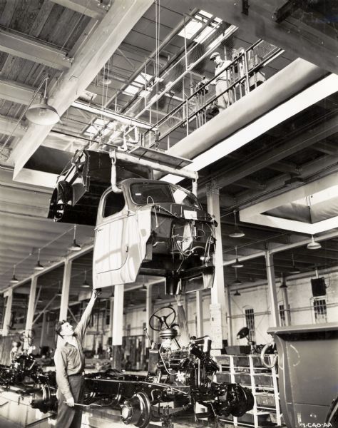 A factory worker standing by a truck cab as it is lowered onto the assembly line at International Harvester's Springfield Works. Another man near a railing is standing above looking down at the truck cab.