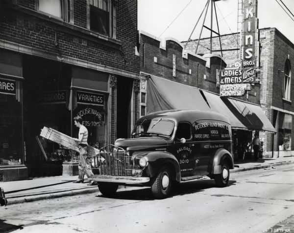 An International KB-3 truck used by the Petersen-Lund Paint Company is parked along the curb in front of a block of commercial buildings. A man on the sidewalk is walking away from the truck carrying a ladder. The original caption reads: "Petersen-Lund Paint Co. 4023 Irv. Pk. Road uses a KB-3 for delivering paint." The text on the side of the truck reads,:"Painters Supplies, Nordica Finishes, Wallpaper."