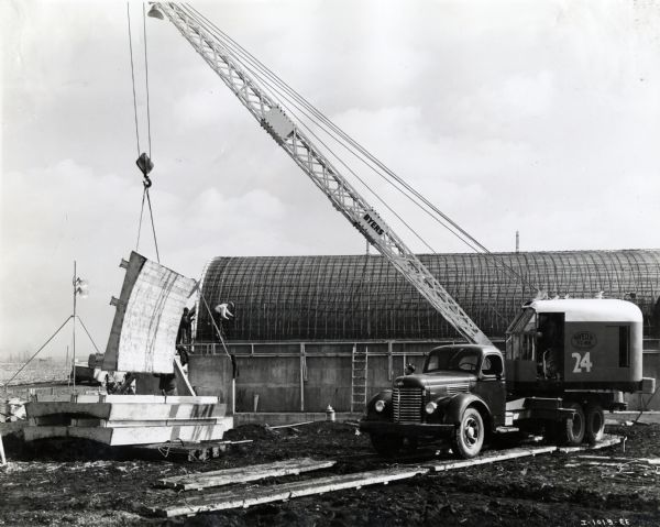An International K-8 truck and a Byers crane are used to lift a piece of wood in the construction of a U.S. Army building. The original caption reads: "National Defense Pictures - Motor Truck Field Scenes; K-8 owned by Bates & Rogers Construction Co., of Chicago, working on U.S. Army Ordinance Plant Construction. Truck has special Tucson extra rear axle & Byers T-65 truck crane. Used for placing and removing concrete forms."