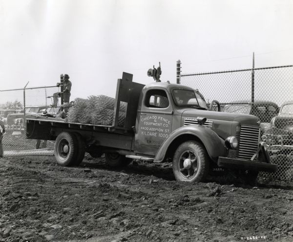 Coils of chain-link fencing are piled on the back of a parked International K-5 truck used by the Chicago Fence & Equipment Company. Behind the truck men are erecting a fence and there are automobiles parked in the background. The original caption reads: "K-5 M.T. owned by the Chicago Fence and Equip Co. Chicago. This Co. is constructing a fence around the new Buick plant at Melrose Park, Ill. This plant will manufacture airplane engines for the govt. & covers 140 acres of land."