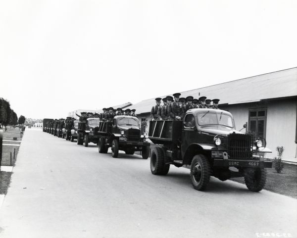 Groups of uniformed Marines await transportation to a rifle range on the beds of International M-3-4 trucks parked in a row along the side of a road. There is a manicured lawn with paths and trees on the left, and on the right in the background are long buildings. The original caption reads, "Fleet of International trucks of various types loaded with Marines at Marine Corps Base, San Diego, ready for transport to rifle range. Truck in foreground is International M-3-4 1 1/2-ton 4-wheel-drive unit with standard cab."