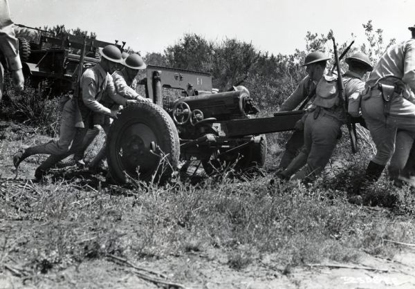 A group of uniformed Marines placing a gun in position an area of brush. An International truck is in the background. The original caption reads: "Marines placing 75-mm. gun in position for firing.  Gun has just been unhitched from truck in left background."