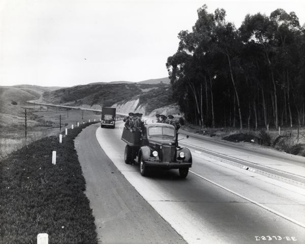 A group of Marines rides on the bed of an International truck as it rolls along a highway.  The original caption reads, "Photograph made on highway north of San Diego near La Jolla. Truck in foreground is Model K-7 with dump body transporting Marines to rifle range, Marine Corps truck in background is Model D-60 with semi-trailer used to haul sea hags and other equipment."