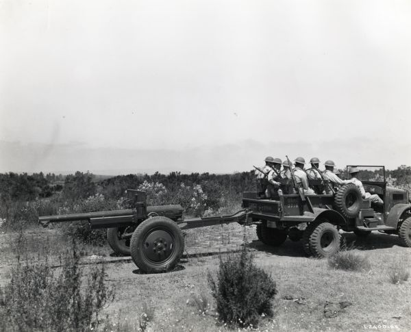 A group of uniformed men ride in the back of an International vehicle which is towing a firearm. The original caption reads: "M-2-4 truck and 75-mm. gun, artillery range, Camp Elliott."