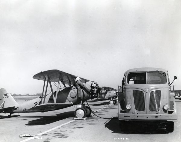 An International KR-11 truck used for refueling parks next to a line of airplanes on a runway. There is a man standing on one of the planes holding a hose attached to the truck. The original caption reads, "One of many International Model KR-11 Cab-over-engine tank trucks owned by the United States Navy and used in refueling Navy planes. International trucks of many models and body styles are in the service of the naval forces. The unit shown has two 750 gallon gasoline compartments and a 100 gallon oil compartment and is equipped with individual power pumps operated by the truck engine."