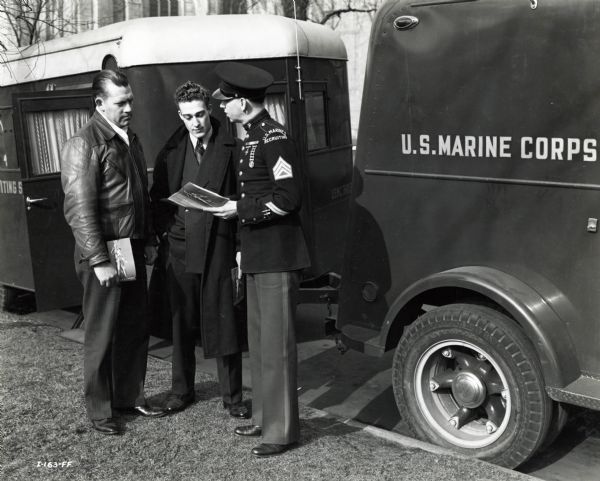 A uniformed Marine stands beside an International U.S. Marine Corps recruiting trailer to speak to two men. The original caption describes the interior features of the marine recruiting trailer as being an "oil heating unit at right, tables and seats which are easily and quickly converted into comfortable beds; the cabinets below the rear window contain gasoline cooking stove, sink, storage batteries, generator, etc."