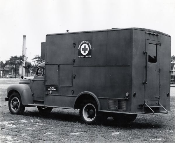 Side view of an International truck used as a mobile disaster unit by the Detroit Chapter of the American Red Cross. The truck is parked in what appears to be a field or park, and a factory building, chimney, houses, and a pedestrian is in the background.