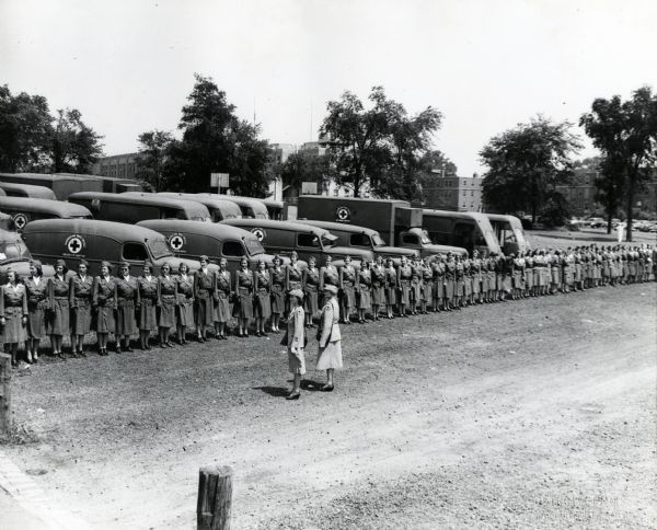 Elevated view of a line of uniformed women employed by the American Red Cross Disaster Unit standing near rows of parked International trucks.