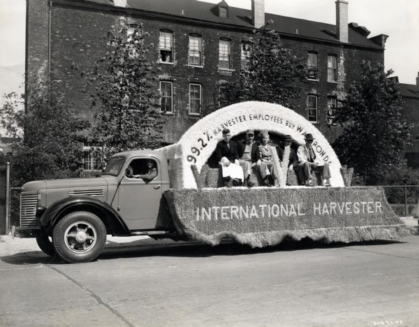 Men and women ride on an International float in the United Nations Parade. The text on the float reads: "99.2% Harvester Employees Buy War Bonds."