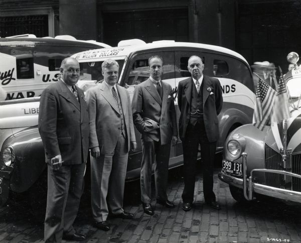 Four men, including Fowler McCormick (third from left), stand in front of the "Liberty Fleet" of International trucks decorated with United States flags. Four of the men appear to be armed guards wearing uniforms with hats. The original caption reads: "'Liberty Fleet' Brinks Express Company."