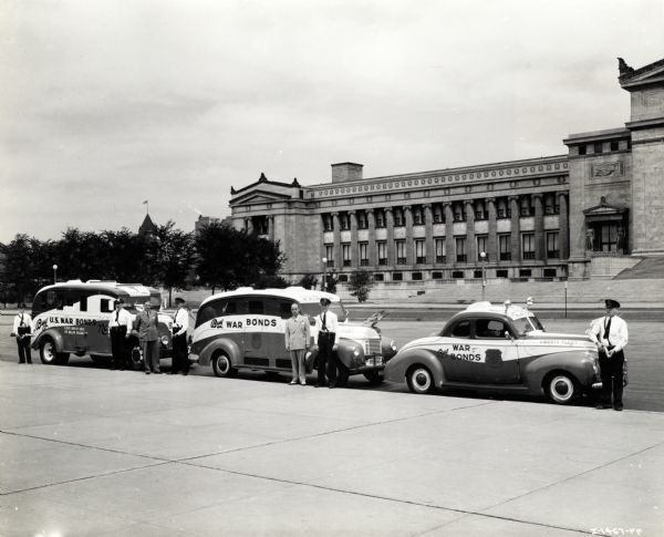 Men stand on the curb beside International trucks used in the Brinks Express Company's "Liberty Fleet." The Field Museum building is in the background across the street. The text on the automobiles reads: "Buy War Bonds" and "Illinois Monthly Quota 85 Million Dollars."