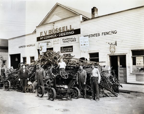 Six men stand outside around a pile of scrap metal in front of the F.V. Russell McCormick-Deering dealership. The metal was collected for a wartime scrap drive. The original caption reads: "This picture shows the results of three days of scrap gathering by F.V. Russell, IHC dealer, Lowell, Indiana. Last Thursday Mr. Russell and his men began. They went out with trucks to farms and demanded the scrap that farmers declared before they had no time to collect. Before this week is out the pile may be much bigger. Mr. Russell piled the scrap in the street right in front of his place of business. Men showing are left to right as follows: Harold Sorensen, F.V. Russell, J.L. McKinney, Lewis Starr, Junior Nichols, and W.E. Bruce."