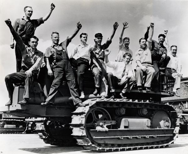 A group of men from Tractor Works who joined the "Harvester Battalion" sit on a military vehicle and raise their arms for a group portrait.