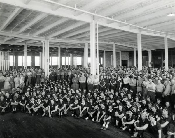 Factory workers sitting and standing for a group portrait at International Harvester's St. Paul Works. Uniformed women are sitting in the foreground, and American flags are hanging from the ceiling.