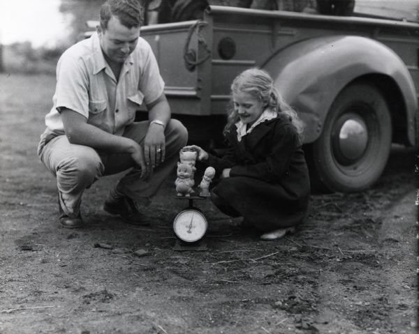 Annabel Nelson places three rubber toys on a scale, presumably to donate them to the war effort, while F.E. Williams looks on.  The two crouch on the ground near the rear of a truck. The original caption reads, "Little Annabel Nelson of near Dayton smiles as she turns in her rubber toys to Blockman F.E. Williams."