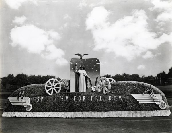 A woman dressed as the Statue of Liberty poses on a General Electric parade float. The sign on the float reads: "Speed 'em for Freedom." The original caption reads: "General Electric float shown in month of July parade, which won first prize. The float was built on one of our model K-7 chassis."