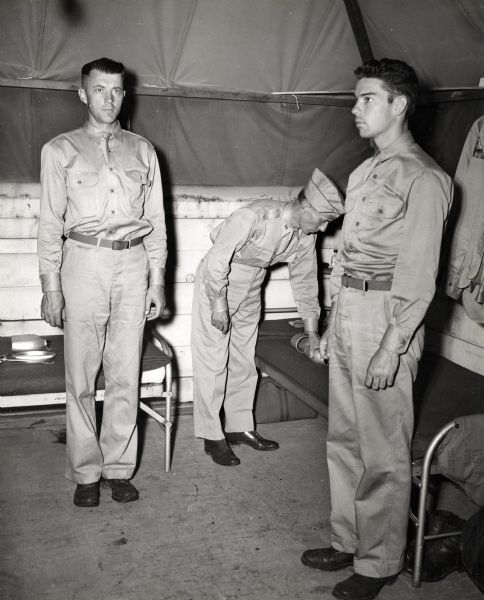 Major Paul H. Wood, center, inspects cots in a tent while two of the tent occupants, former employees of the International Harvester Company, stand at attention on either side of him. The men likely belonged to the "Harvester Battalion," and may have been stationed at Camp Perry, Ohio for training. The original caption reads: "Major Paul H. Wood (center) takes a morning trip through the camp to see that things are in order. The tent occupants standing at attention are Sgt. James R. Chrisman (left), former dealer, Richmond, Ky., and Joseph J. Stanton, Pvt.F.C., formerly of the Philadelphia (f.e.) branch.
