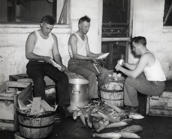 Three soldiers, formerly International Harvester employees and dealers, sitting on wooden crates to shuck ears of corn near a doorway. The men likely belonged to the "Harvester Battalion," and may have been stationed at Camp Perry, Ohio for training. The original caption reads: "AN OLD STORY IN THE ARMY. Not spuds this time but their equivalent in corn. Left to right: Fred M. Bourke, former dealer, Lewistown, Mont.; Worth A. Stewart, former dealer, Carriso Springs, Tex; Carl C. Moellering, from service shop, Fort Wayne branch."