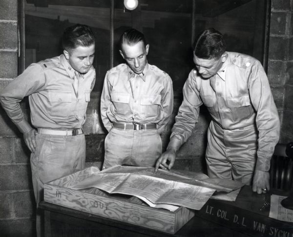 Three uniformed men study a map placed on a desk. The original caption reads: "STUDYING THE BATTALION'S NEXT HOME. Lt. Col. D.L. Van Syckle (right), commanding officer of the "Harvester Battalion," with Major Paul H. Wood (center) and 2nd Lieut. John C. Bond look over a map and details of Camp Campbell, Ky., where the battalion will go after completion of basic training at Camp Perry."