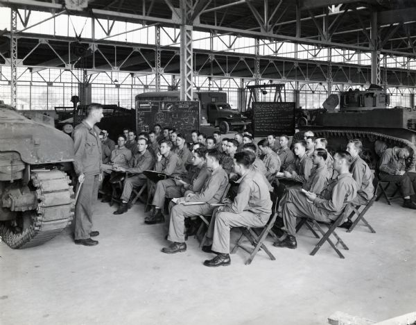 A group of uniformed military men sit in wooden chairs inside a building at Camp Perry as they are instructed on various kinds of machinery. Two chalkboards and various equipment and vehicles are in the background. The men may belong to a group of former International Harvester employees known as the "Harvester Battalion."