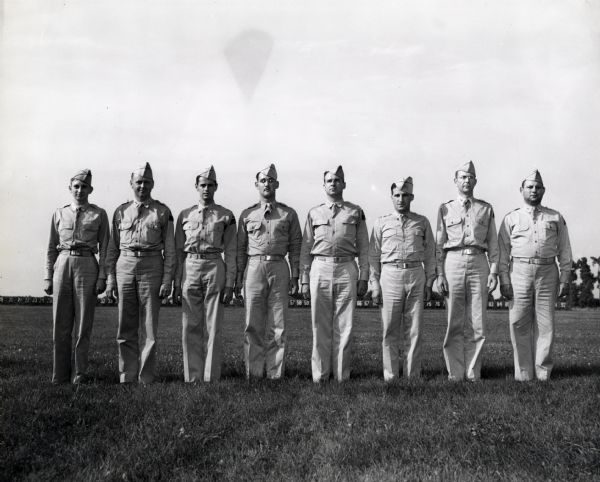 Eight officers assigned to train the "Harvester Battalion" stand in a line in a field for a group portrait. In the background is a row of numbers. The men may be stationed at Camp Perry, Ohio. The original caption reads: "IN CHARGE OF BASIC TRAINING. These experienced officers were attached to the "Harvester Battalion" to give the men basic training. They are (left to right): 1st Lieut. Robert Bennet, Capt. George L. Johnson, Capt. Joe H. Serkowich, Capt. Arthur F. DeLong, Major Leland K. Warrick, Capt. Louis Ebert, Capt. Ura Morgan, 1st Lieut. Emmanuel Goldstein."